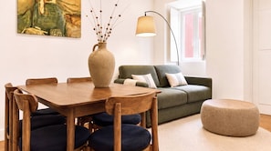 A dining area with a wooden table and chairs, a vase with dried branches, a green sofa with cushions, a floor lamp, and a window with a view. - VIVA Janelas Verdes Residence (Lisboa)
