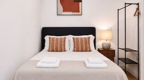 A neatly made bed with white linens and two decorative pillows, a bedside table with a lamp, and a wall-mounted shelf with a hanger. - VIVA Janelas Verdes Residence (Lisboa)