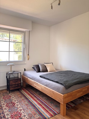Ein Schlafzimmer mit einem Bett, einem Nachttisch, einer Lampe, einem Fenster und einem gemusterten Teppich.