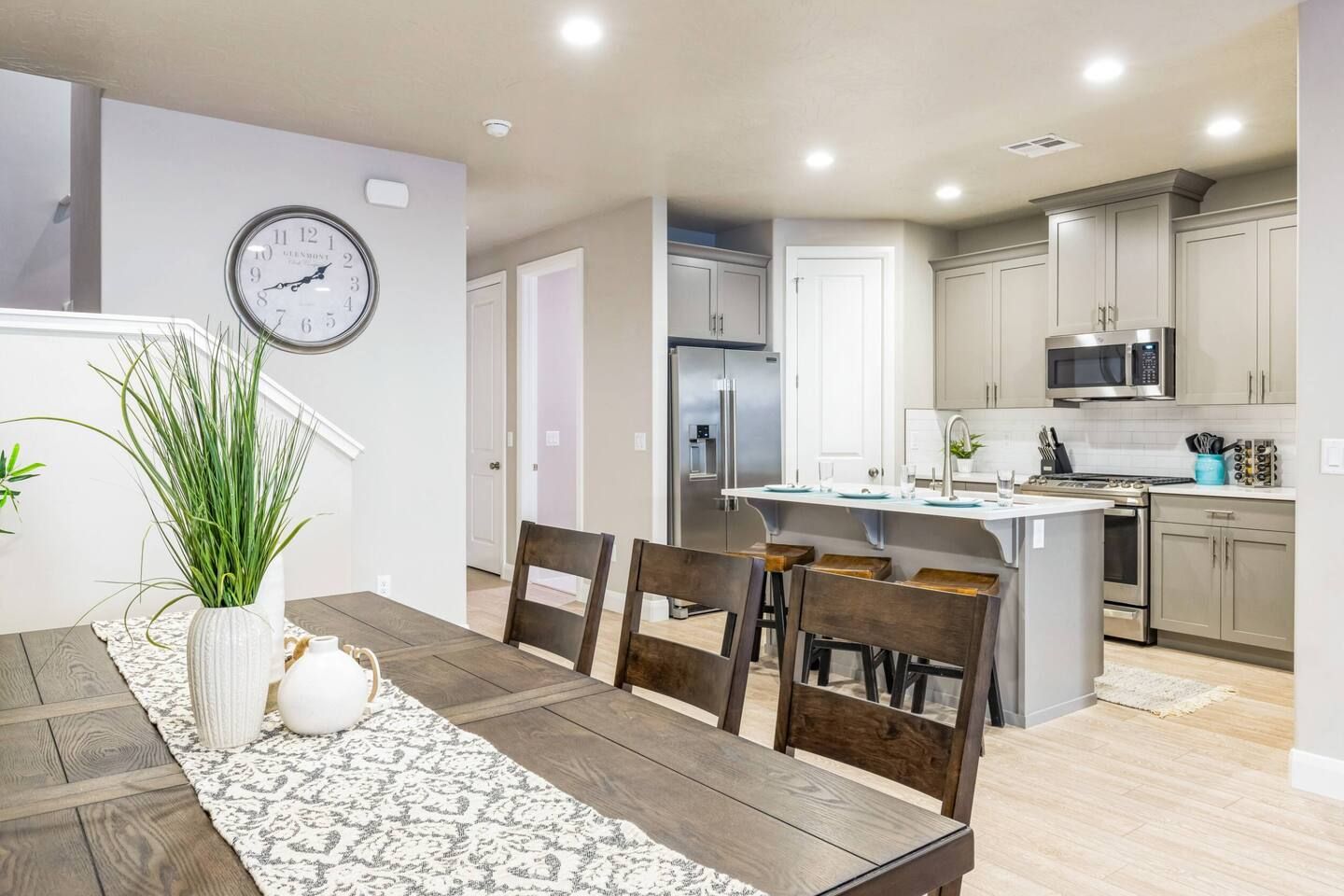 A modern kitchen with stainless steel appliances, a dining area with a wooden table and chairs, and a clock on the wall.