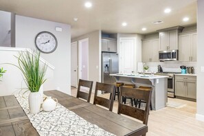 A modern kitchen with stainless steel appliances, a dining area with a wooden table and chairs, and a clock on the wall. - Heated Pool & Pickleball - Beautiful 3BR Home Near Zion (Washington)