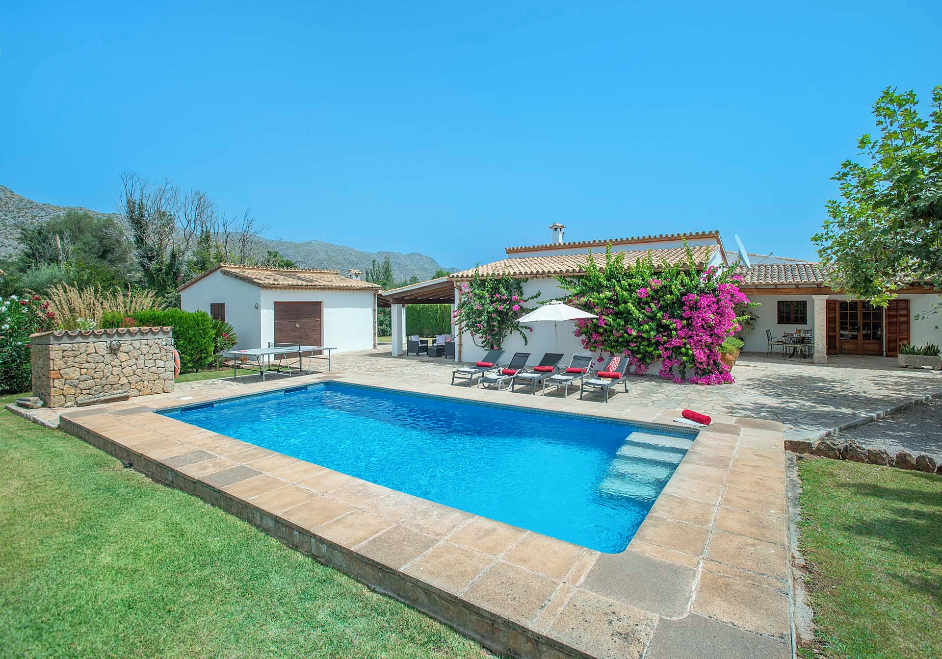 Una piscina con zona de patio, mobiliario exterior y un edificio blanco con tejado de tejas.