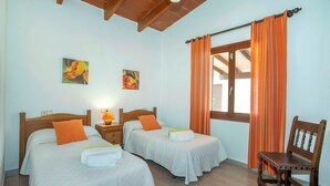 A room with two beds, a nightstand with a lamp, a wooden chair, and a window with curtains. - Countryside Retreat in La Font, A/C, Private Pool, Large Garden - 15mins Walk to Old Town Pollenca! (Pollença)