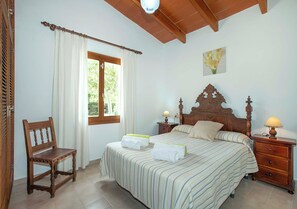 A bedroom with a large bed, a wooden chair, a nightstand, and a window with curtains. - Countryside Retreat in La Font, A/C, Private Pool, Large Garden - 15mins Walk to Old Town Pollenca! (Pollença)