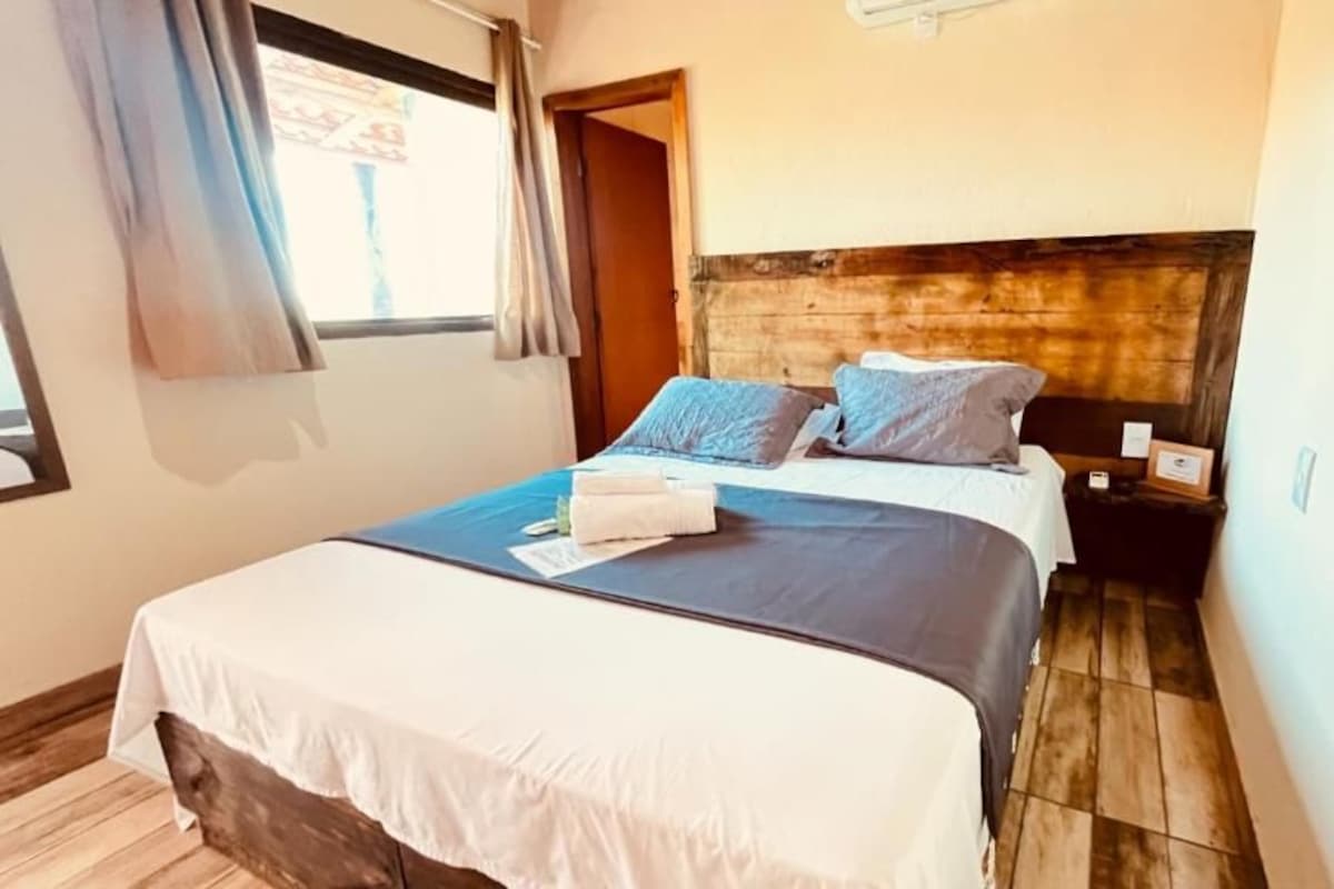 A double bed with a white and dark-colored bedspread, two pillows, a bedside table with a lamp, and a window with curtains.