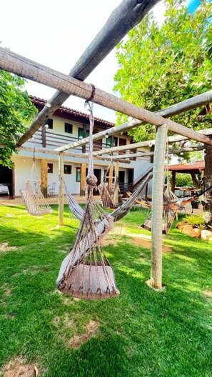 A hammock under a wooden structure, a building with a red roof, and a grassy area. - Pousada Flor de Pequi (Santana do Riacho)