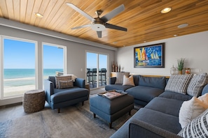 Ein geräumiges Wohnzimmer mit großem Fenster mit Blick auf den Strand, einem Deckenventilator und einem Bild an der Wand.