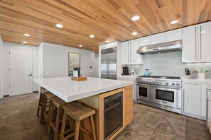 A modern kitchen with a central island, wooden stools, a stainless steel refrigerator, a gas stove, and white cabinetry. - Oceanfront 3BR Condo • Private Beach Access • Oceanside Pier Views (Oceanside)