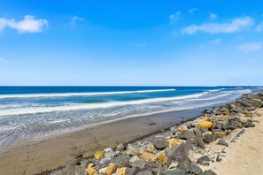 Ein Strand mit Wellen, ein Steg in der Ferne und eine felsige Küstenlinie.