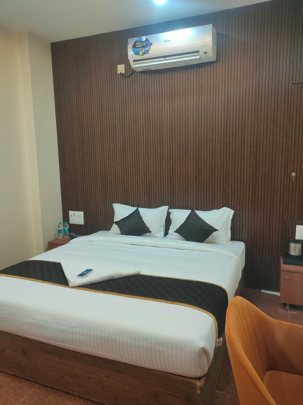 Quarto de hotel com cama, cabeceira de madeira, ar condicionado, mesa de cabeceira com telefone e garrafas de água, e uma cadeira de madeira.