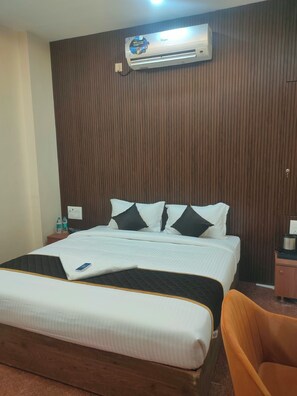 Una habitación de hotel con una cama, cabecera de madera, unidad de aire acondicionado, mesita de noche con un teléfono y botellas de agua, y una silla de madera.