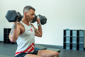 Eine Person trainiert mit Kurzhanteln in einem Fitnessstudio, im Hintergrund sind eine Bank und Stauraumelemente zu sehen.