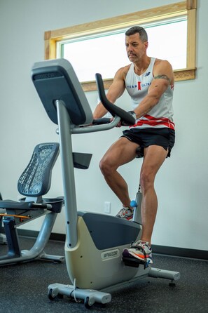 A person using a stationary bike in a gym with a window and a chair in the background. - The Hotel Cabin - Bold Americana Suite - Pool - Hot Tub - Gym - Bar - Lake (Crystal River)