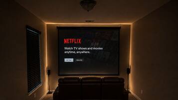 A home theater room with a large screen displaying a Netflix interface, leather reclining seats, and a carpeted floor.