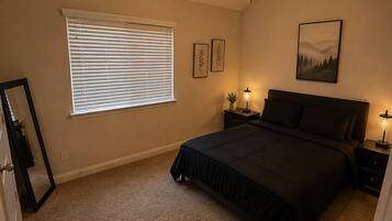 Een slaapkamer met een bed, een plafondventilator, een raam met jaloezieën, bedlampen en een kunstwerk in een lijst aan de muur.