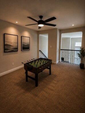 Een kamer met een tafelvoetbaltafel, een plafondventilator en twee ingelijste foto's aan de muur.