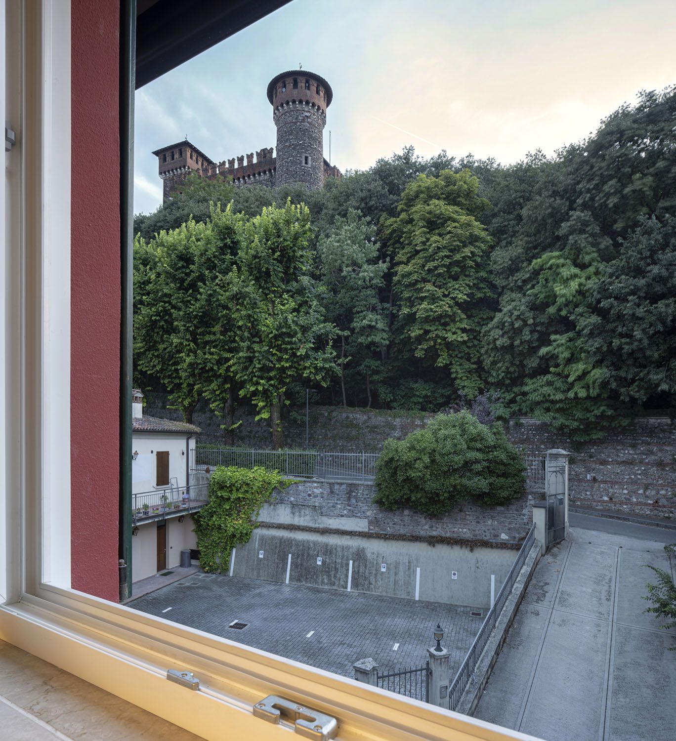 On aperçoit, depuis une fenêtre donnant sur une zone pavée bordée d’une clôture et surmontée d’un petit bâtiment, le sommet d’une tour de château et la végétation luxuriante qui l’entoure.
