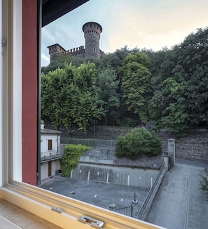 On aperçoit, depuis une fenêtre donnant sur une zone pavée bordée d’une clôture et surmontée d’un petit bâtiment, le sommet d’une tour de château et la végétation luxuriante qui l’entoure.