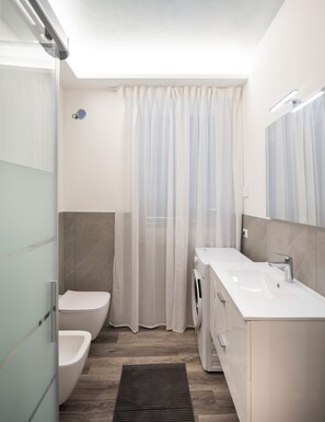 Ein modernes Badezimmer mit Glasdusche, Toilette, Bidet, Waschmaschine und Waschbecken mit Spiegel.