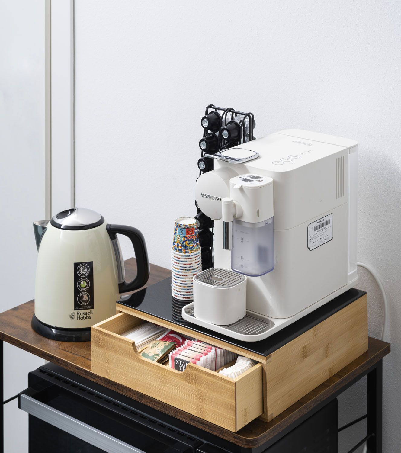 Una cafetera, un hervidor y un cajón con cápsulas de café sobre una mesa de madera.