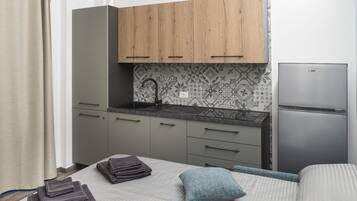 A compact bedroom with a bed, pillows, a neatly folded towel, a small bedside table, and a kitchenette area with a sink, cabinets, and a refrigerator.