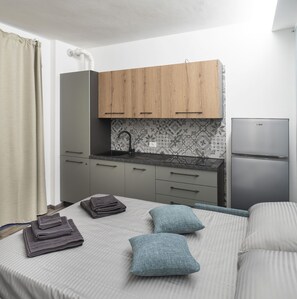 Ein gemütliches Schlafzimmer mit Bett, Kissen, einem ordentlich zusammengefalteten Handtuch, einem kleinen Nachttisch und einer Küchenzeile mit Spüle, Schränken und einem Kühlschrank.