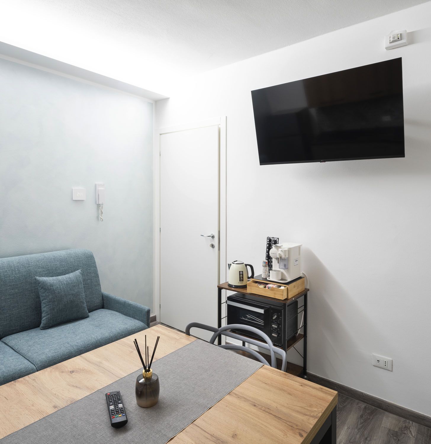 Una sala de estar moderna con un sofá, una mesa de centro, un televisor montado en la pared y una pequeña zona de kitchenette.