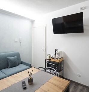 A modern living room with a sofa, a coffee table, a TV mounted on the wall, and a small kitchenette area.