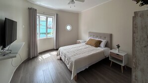 Een slaapkamer met een bed, een televisie, een nachtkastje, een ventilator in het plafond en een raam met gordijnen.