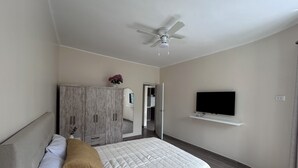 Een slaapkamer met een bed, een kledingkast, een plafondventilator, een televisie op een standaard en een deur die naar een andere kamer leidt.
