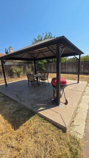 Eine überdachte Terrasse im Freien mit einem Grill, einem Tisch mit Stühlen und einem Holzzaun im Hintergrund.