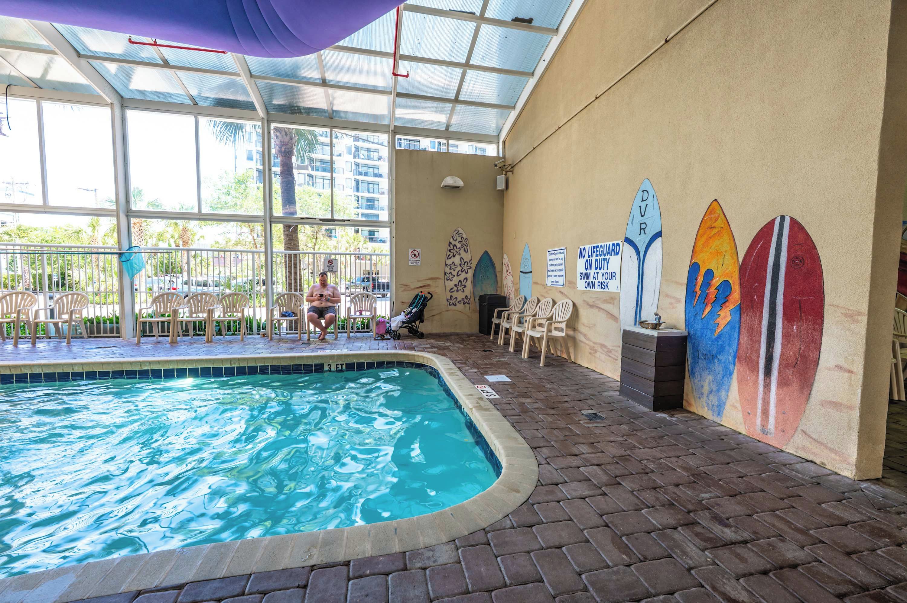 An indoor swimming pool with a tiled floor, a glass ceiling, and a mural of surfboards on the wall.