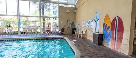 An indoor swimming pool with a tiled floor, a glass ceiling, and a mural of surfboards on the wall.