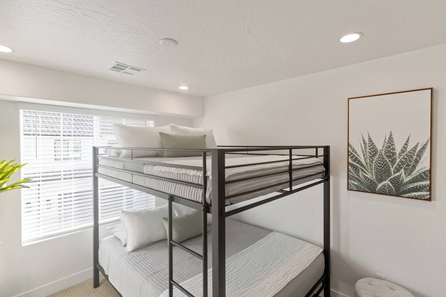 A bunk bed with white bedding, a window with blinds, and a plant on the left.