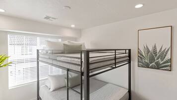 A bunk bed with white bedding, a window with blinds, and a plant on the left.