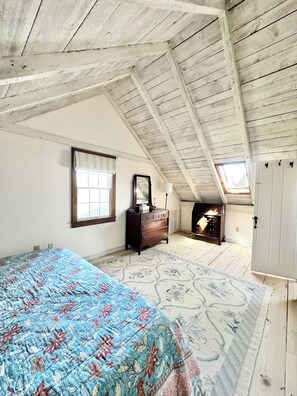 A bedroom with a sloped ceiling, a bed with a floral bedspread, a dresser, a mirror, a window, and a fireplace.
