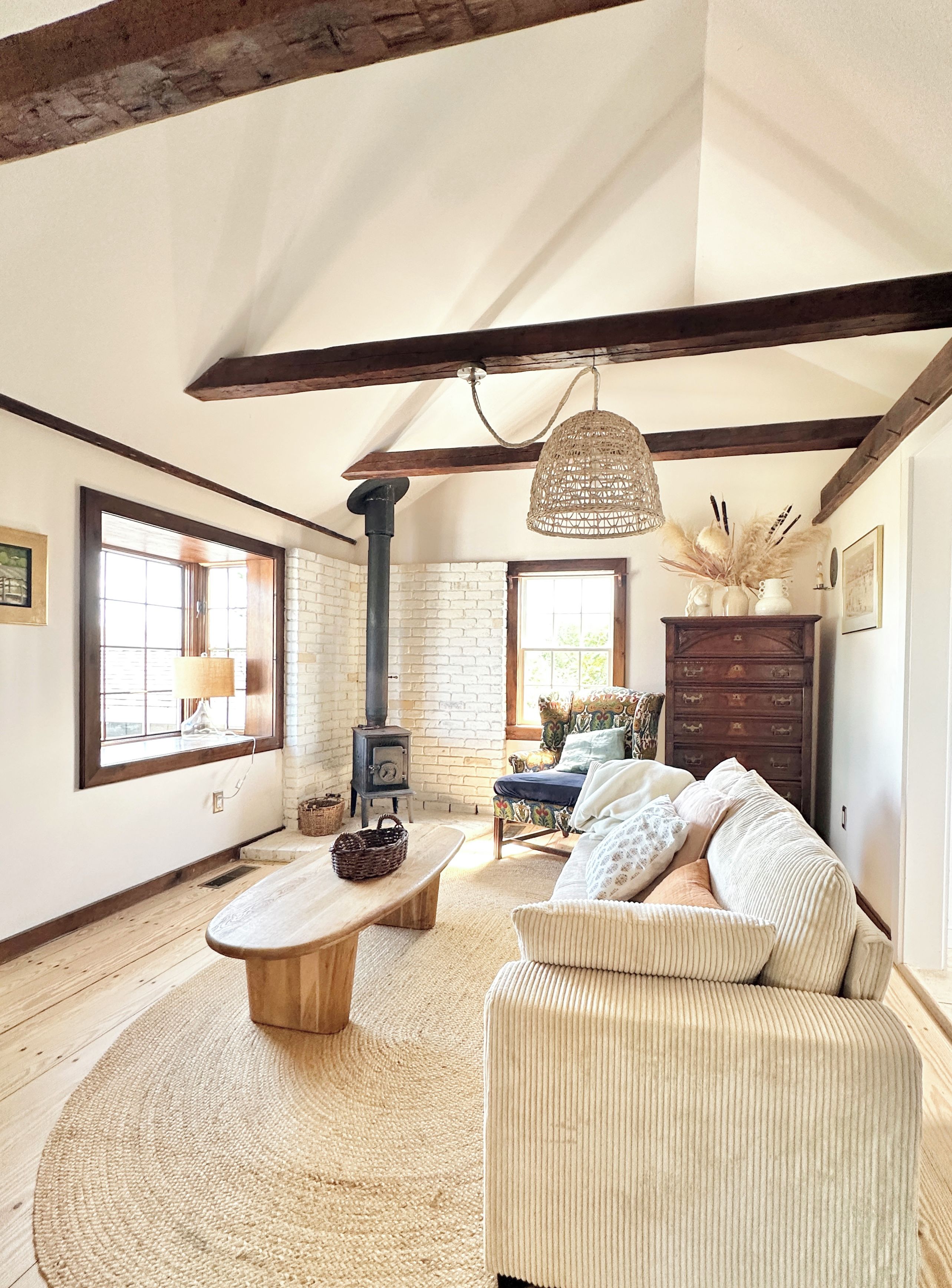 A rustic living room with exposed wooden beams, a stone fireplace, a woven light fixture, and a comfortable sofa.