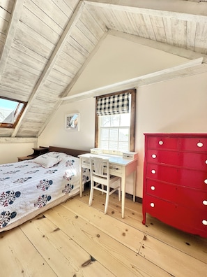 A bedroom with a sloped ceiling, a bed with a floral bedspread, a desk with a chair, a red dresser, and a window with a checkered blind.