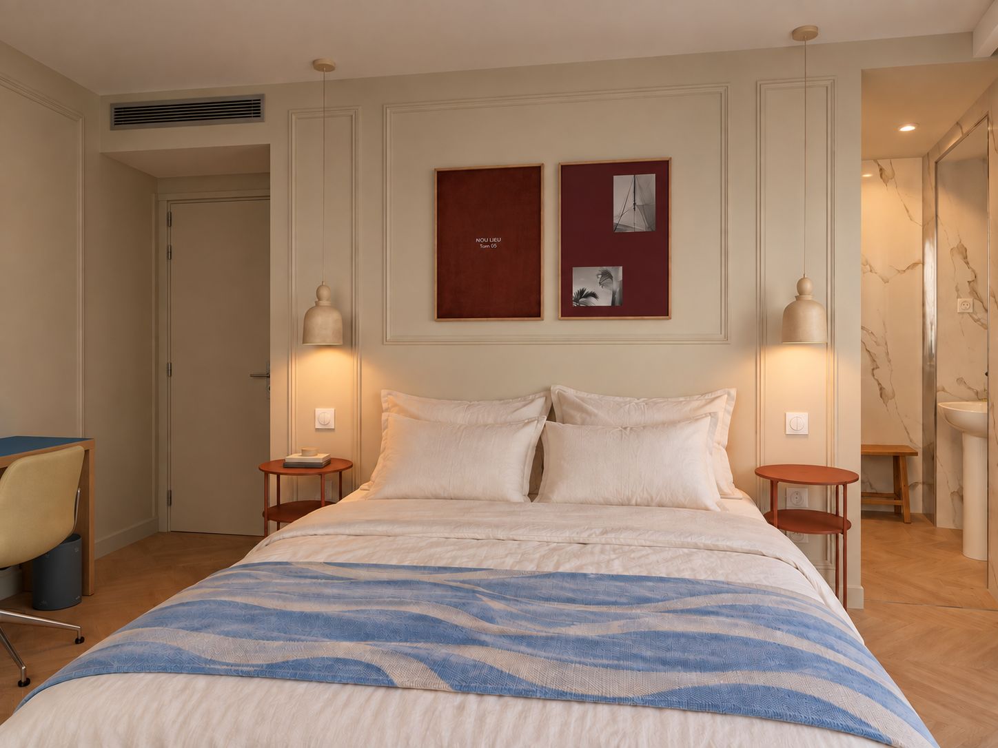 A hotel room with a large bed, two bedside tables, a chair, and a desk. There are two wall-mounted picture frames and a ceiling vent.