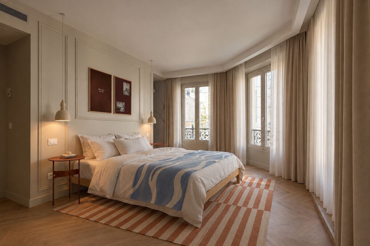 Un dormitorio con una cama grande, una mesita de noche, una silla, una alfombra y una ventana con cortinas.
