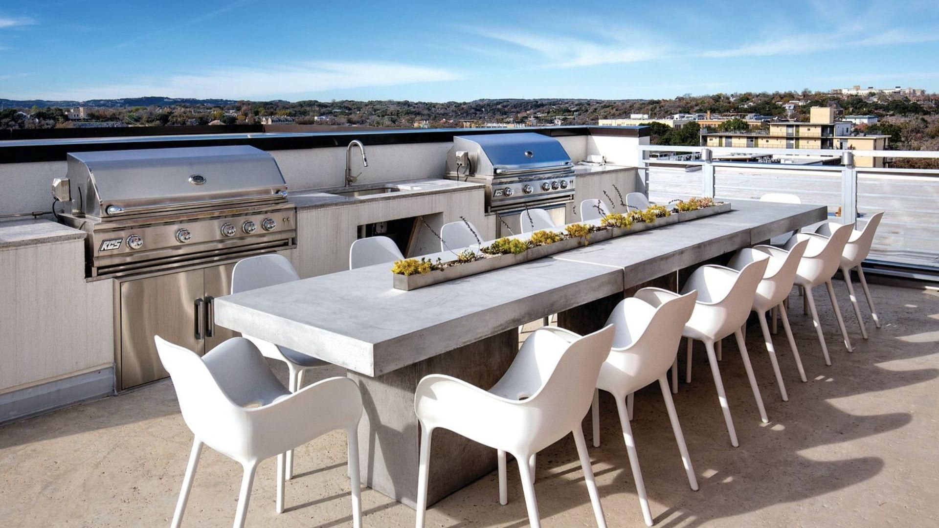 An outdoor patio with a barbecue grill, a long dining table, and white chairs.