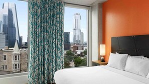 Ein Hotelzimmer mit großem Fenster, einem Bett, einem Nachttisch und Blick auf die Stadtlandschaft.