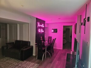 Ein Wohnzimmer mit Essbereich, einer schwarzen Ledersofa, einem Glastisch und Stühlen. An der Wand hängt ein Neon-Schild und rechts befindet sich ein Fernseher.