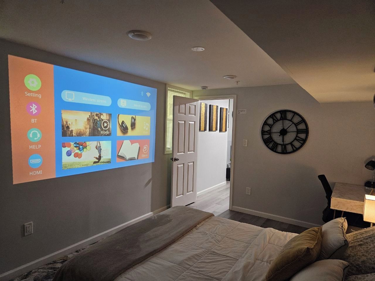 Un dormitorio con una cama, un reloj en la pared y una pantalla de proyección mostrando diversos iconos.