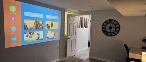 Un dormitorio con una cama, un reloj en la pared y una pantalla de proyección mostrando diversos iconos.