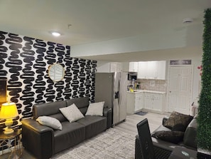 Ein modernes Wohnzimmer mit einer Couch, einem kleinen Tisch mit Lampe und einer Küchenzeile im Hintergrund.