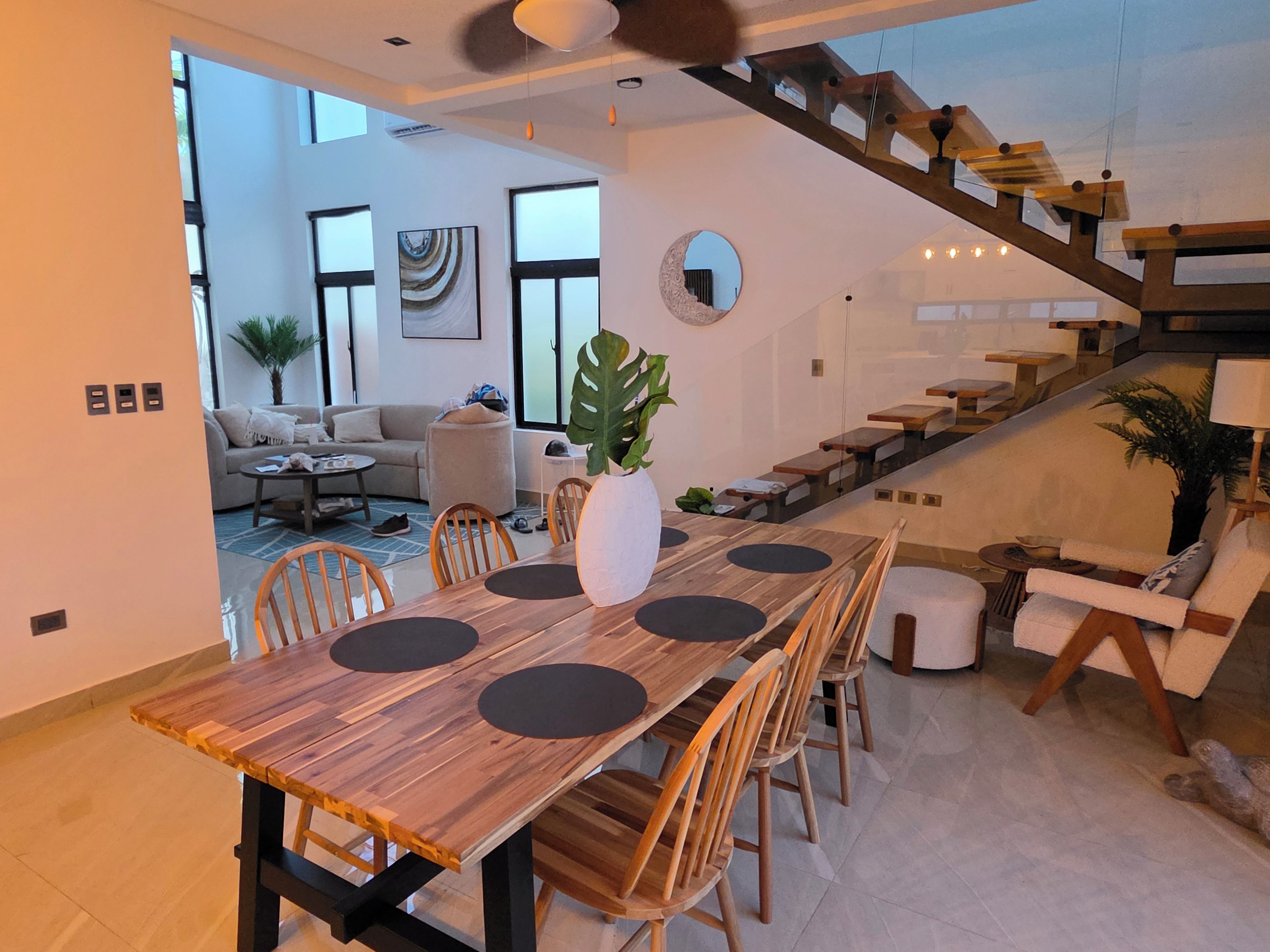A modern dining area with a wooden table and chairs, a large plant centerpiece, and a staircase leading to an upper level.