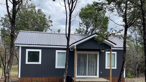 A small, dark-colored cabin with a metal roof, a front door, and a window on the side. - "The Ft Worth" Romantic Tiny Home Near Wineries (Weatherford)