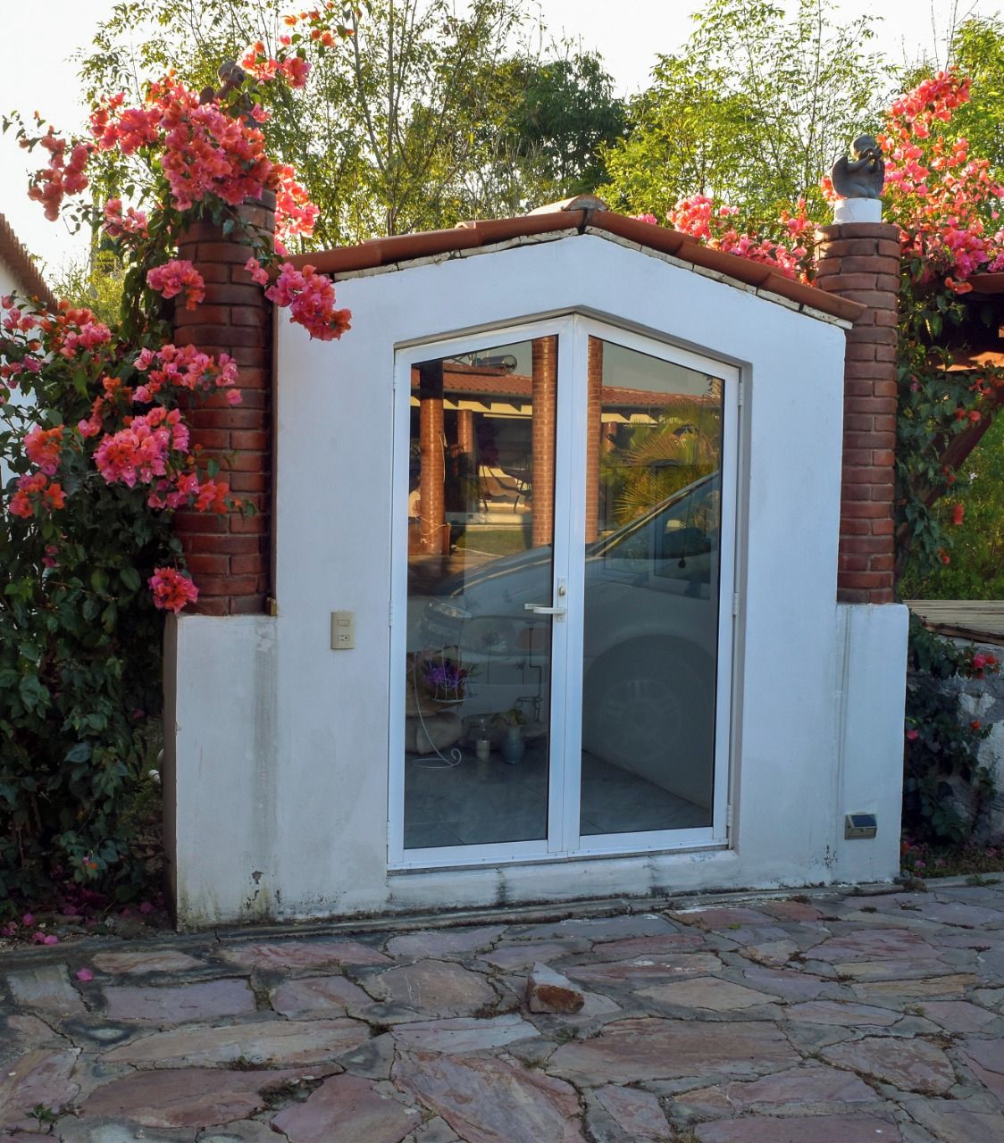 Un petit bâtiment blanc, de plain-pied, doté d’une porte en verre et de cheminées en briques, entouré de plantes à fleurs roses.