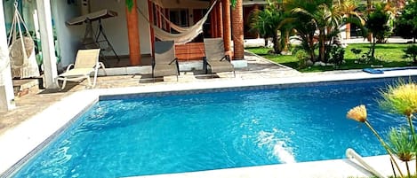 A swimming pool with lounge chairs and a hammock, surrounded by greenery and a building with a tiled roof.
